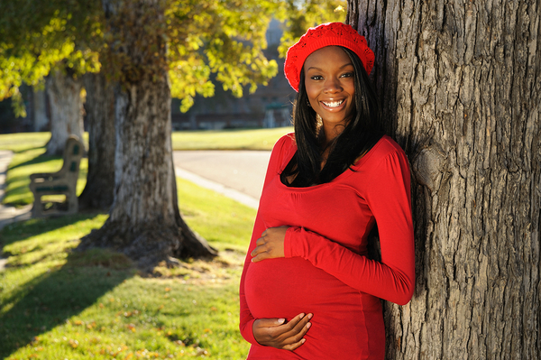 Pregnant woman in red dress and red beret leaning against tree trunk, smiling at camera
