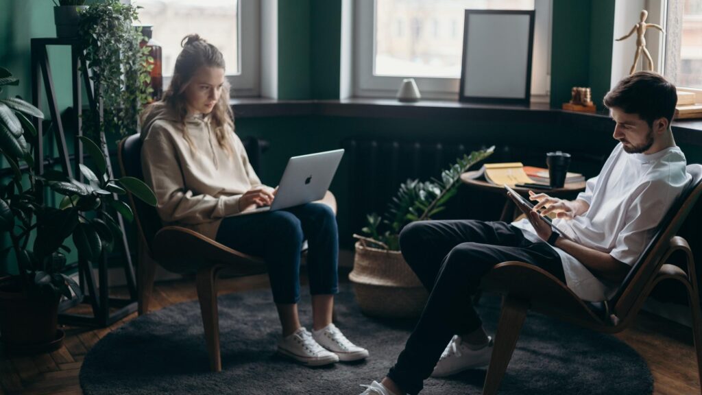 Woman and man sitting in office chairs facing each other, woman using laptop, man writing on clipboard, surrounded by plants and windows