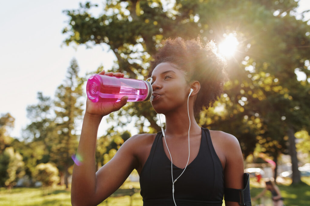 Woman drinking from pink water bottle while exercising outdoors in a park with earbuds in