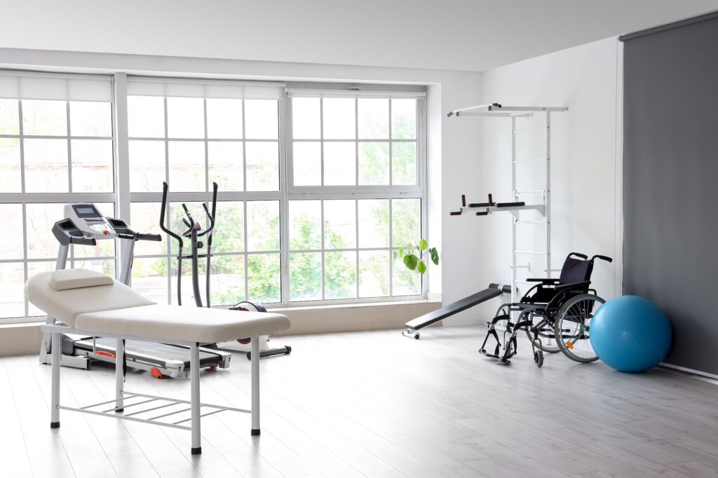 Modern rehabilitation clinic with exercise equipment including treadmill, stationary bike, treatment table, wheelchair, exercise ball, and large windows overlooking greenery.