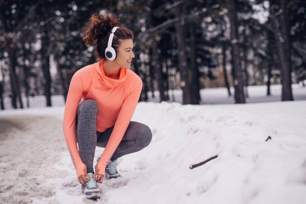 Woman in orange long-sleeve shirt and gray leggings stretches while wearing white headphones in snowy forest setting