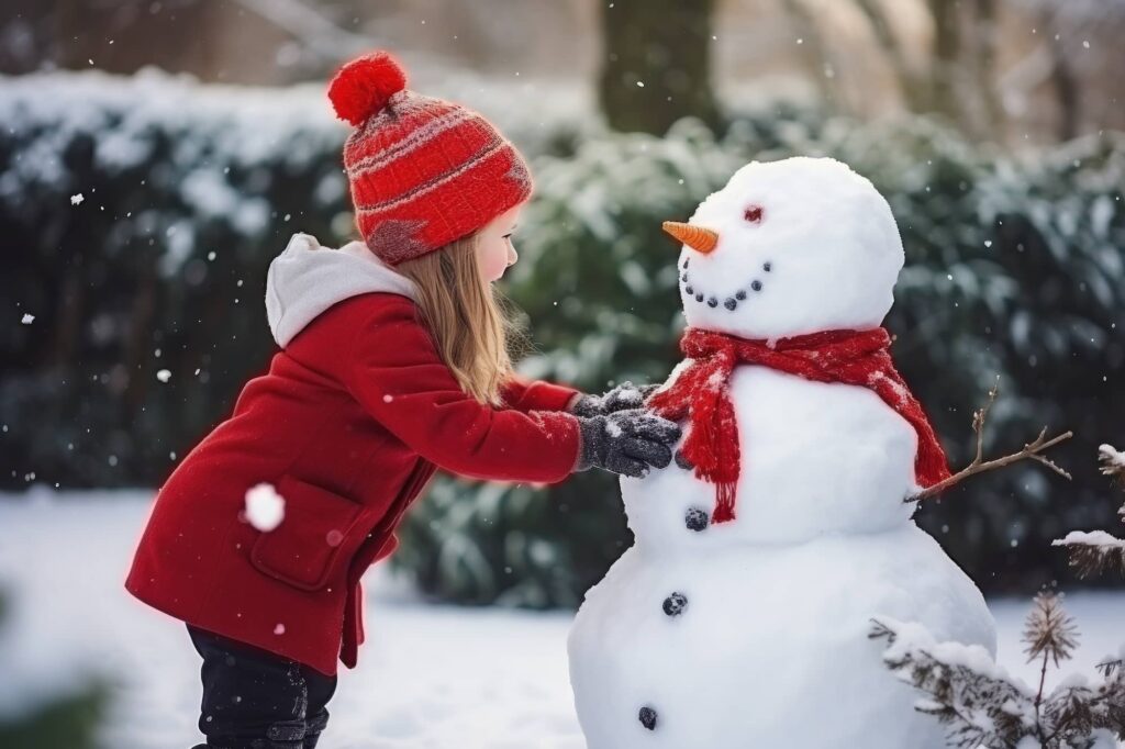 Child in red winter coat and knit hat touching a snowman wearing a red scarf in falling snow